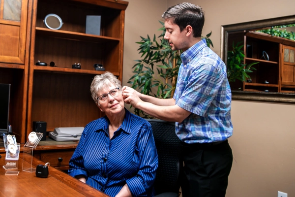 An audiologist is fitting a hearing aid on an elderly woman seated at a desk in an office setting.